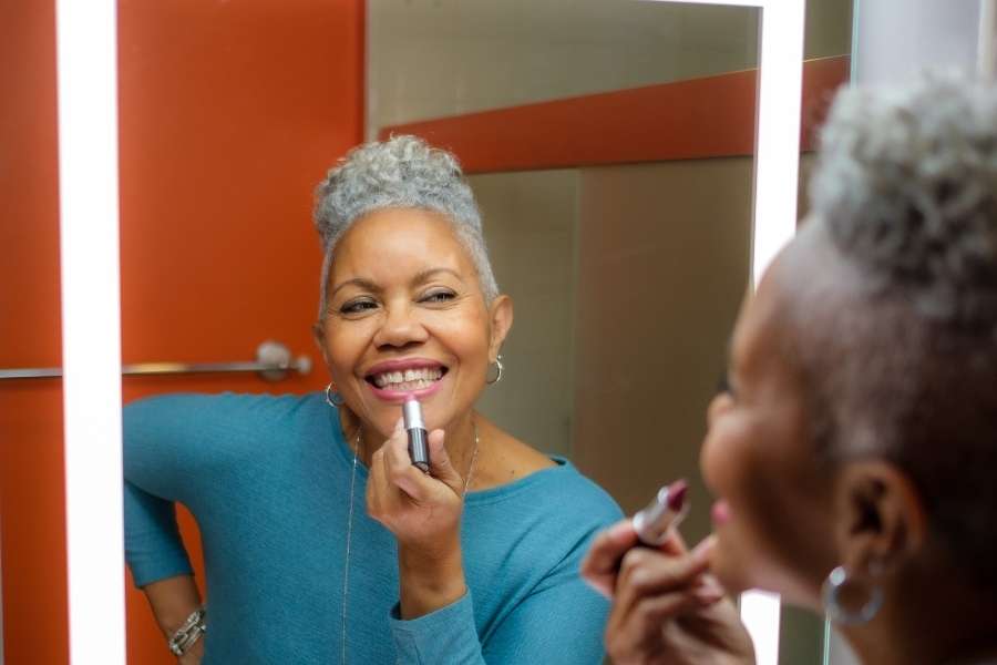Une femme senior aux cheveux gris se maquille les lèvres devant un miroir lumineux.
